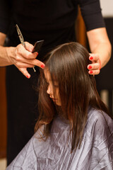 Fototapeta premium Portrait of a young girl getting her hair cut in home. Mom's hand with hairdressing scissors. Little child sits patiently whilst having her hair cut.