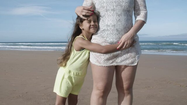 Mom and cute black haired girl hugging while standing on ocean beach. Cropper shot. Family summer vacation concept