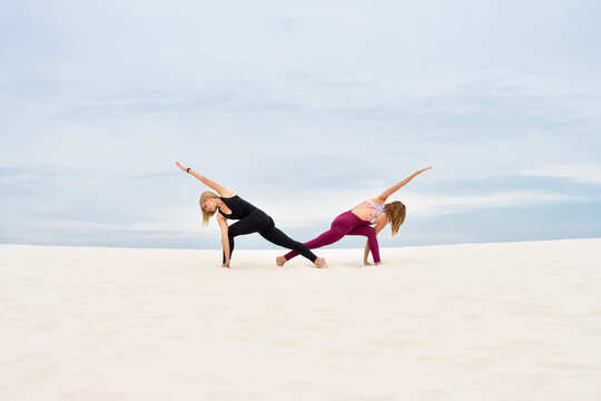 Two beautiful young women performing yoga pose parivrita parshvakonasana together on the beach