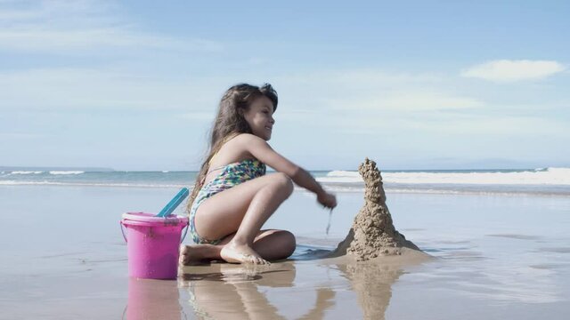 Happy cute little girl building sandcastle on beach, playing with wet sand near toy bucket and shovel. Childhood and outdoor activity concept