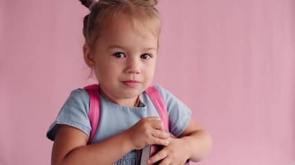 childhood, school, education, upbringing, science concept - close-up little blonde caucasian slavic girl in with backpack holding book and showing discontent and disagreement on solid pink background