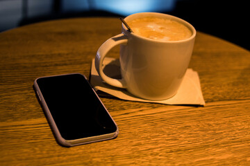 Fototapeta na wymiar White cup of coffee on a table in a coffee shop with a phone next to it