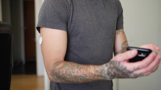 A tattooed young man with diabetes uses his phone to check his blood glucose levels with glucose reading sensor on his arm and then watches the info on the phone.