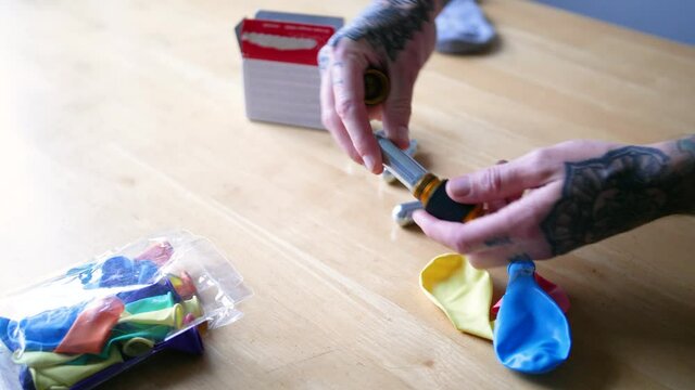 A young man with tattoos is blowing up a colorful balloon with nitrous oxide (N2O) through a gas cracker device. He is using a sock/wool cloth to protect his hands from the intense, cold gas.