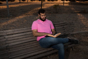 Caucasian man reading a book in the park while wearing a pink shirt and jeans