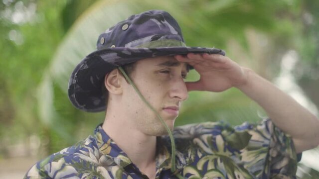 Young Male Tropical Jungle Explorer scans the horizon as he explores a deserted beach. Wearing Hawaiian Shirt and Camouflage Wide Brim Hat