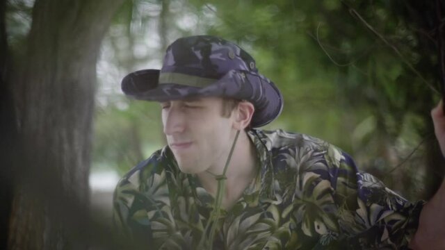 Young white man explores tropical jungle. Wearing Tilly hat and Hawaiian shirt. Scans the horizon with hand on brim