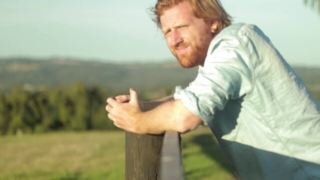 A young farmer in a blue shirt rests on a wooden fence post and looks out across the country in slow motion.
