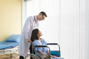 Young Doctor talking to woman in wheelchair after an operation. Smiling doctor looking at a female patient on a wheelchair in hospital. Photo of positive doctor and patient on wheelchair.