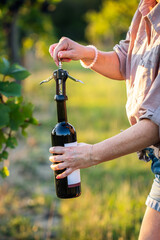 Woman opening red wine bottle by corkscrew at vineyard. Pulling wine cork from bottle outdoors....