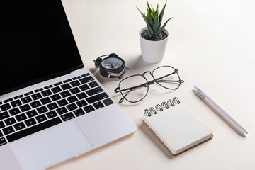 Closeup shot of an open laptop, glasses, a plant, a pen, a notepad, and an alarm clock