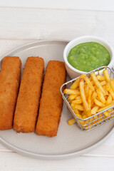 Fish fingers served with chips in a wire basket and mushy peas.  White wood background