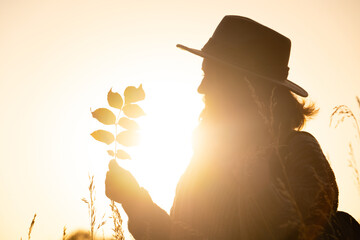 Portrait of a woman wearing a hat, and with a backpack. She holds yellow leaves in her hand at...