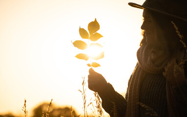 Portrait of a woman wearing a hat, and with a backpack. She holds yellow leaves in her hand at sunset. Autumn, copy space.
