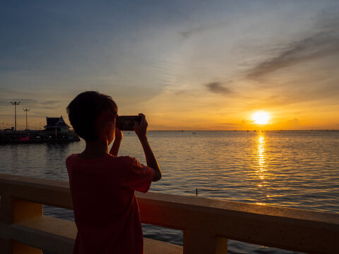 Rear view silhouette of an Asian boy taking a sunset picture with the smartphone.