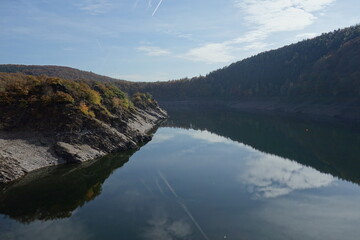 Talsperre umrandet von herbstlichem Wald . Nasser stiller See mit spiegelnder Hügellandschaft . Blass Blauer Himmel mit leichter weißer Bewölkung .