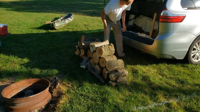 Man taking outfire wood rom his car trunk and setting or placing it or stacking pile next to fire pit at camp site.