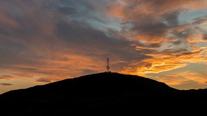 Sunrise behind a hill with a silhouette of transmitter telecommunication antenna on top of hill. Colorful dramatic clouds during sunrise.