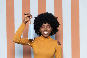 Happy African American woman with curly hairstyle and closed eyes, holding sterile protective medical face mask, posing on striped background outdoors. Health care concept.