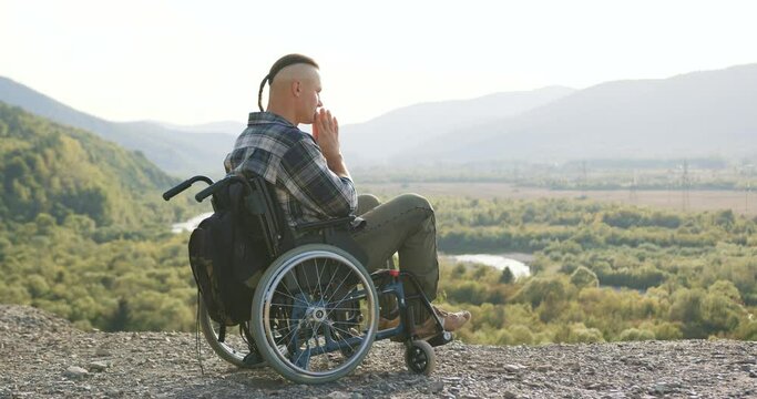Side view of handsome calm concentrated man with little ponytail which sitting in wheelchair on the hill and praying