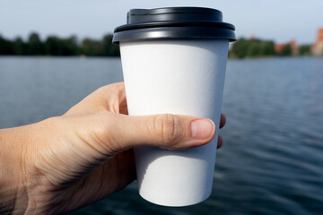 Coffee at the lake. Young woman holds a disposable cup of coffee on on a sunny morning at the beach, lake or river. 