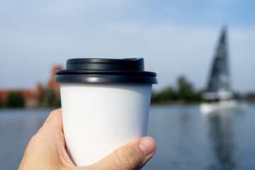 Coffee at the lake. Young woman holds a disposable cup of coffee on on a sunny morning at the beach, lake or river. 
