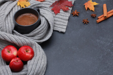 Black fresh coffee mug, red apples, yellow maple leaves, cinnamon, pale grey scarf on dark background. Copy space, high angle. Autumn, fall, thanksgiving, coffee shop menu concept.