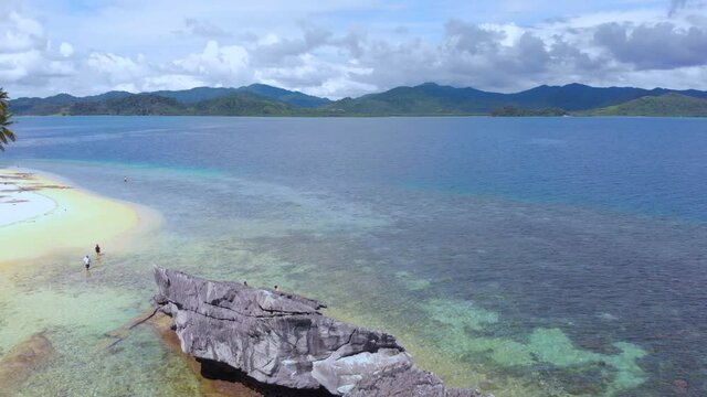 Circling drone shot of large rock formation in the Ocean eventually revealing white sand beach with palm trees on an island with people