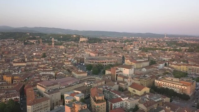 Aerial shot of Verona, Italy at sunset with typical Italian apartment rooftops, street crowds, Roman Colosseum Arena, Adige River, Santuario Madonna di Lourdes, mountains Lessinia Natural Park.