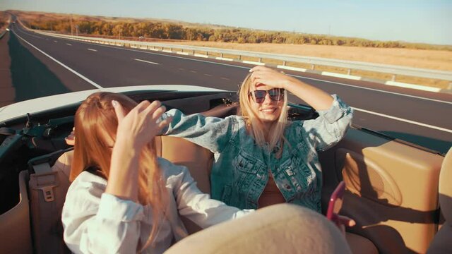 Woman in back seats of convertible take selfies on vacation. Two young women sit in car without roof shooting videos and taking photos on phone, side view