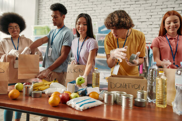 Young male and female volunteers packing food and drinks donation for homeless into paper bags,...