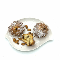 Muffins with raisins on a white porcelain plate in the form of a leaf. Two whole, one broken in half. On a platter, almond grains, crumbs. Isolated on a white background.