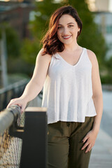 Stunning young woman poses for photo in late summer in elevated urban park - white blouse and green pants - smiling
