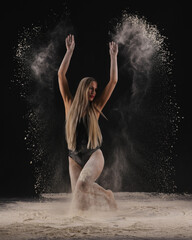 Beautiful sensual fit slim dancer in black body with long blond hair throwing dust, flour, powder on black background.