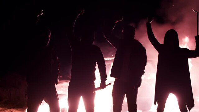 Silhouette of protesting guys with baseball bats and crowbar, raising their hands and shouting slogans, against background of smashed burning car