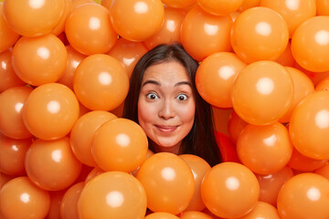 Fototapeta na wymiar Horizontal shot of cheerful surprised brunette young Asian woman surrounded by orange inflated balloons makes decorations for party. Celebration entertainment festive event and holiday concept