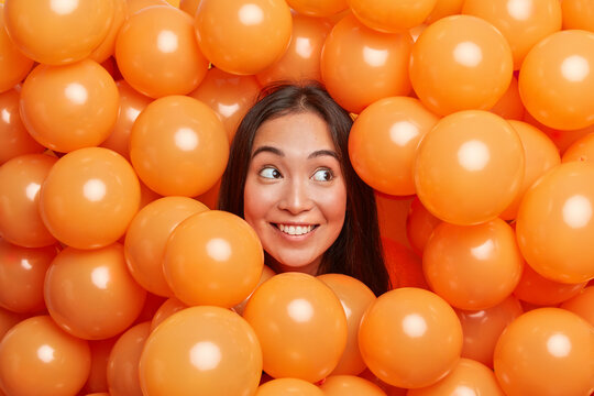 Positive young Asian woman smiles broadly and looks away surrounded by many orange air balloons enjoys birthday party event has long dark hair celebrates something being in good festive mood