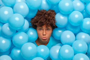 Fototapeta na wymiar Discontent curly haired young woman with miserable gloomy expression has bad mood because of spoiled holiday upset friends didnt come to congratulate her with birthday poses over blue balloons