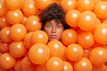 Surprised birthday girl poses over inflated orange party balloons stares bugged eyes expresses...