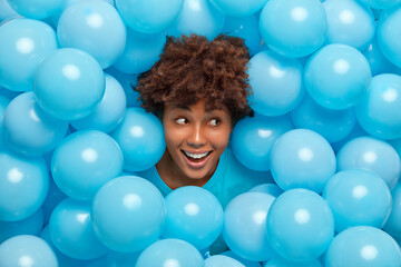 Fototapeta na wymiar Optimistic young African American woman with curly hair and laughs joyfully surrounded by blue balloons enjoys favorite holiday going to have party. Moments of joy and happiness. Great pleasure