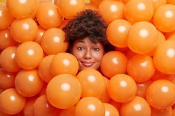 Fototapeta na wymiar Photo of attractive curly haired woman surrounded by inflated balloons smiles gently poses at camera. Happy dark skinned Afro American lady waits for birthday party. Holiday and celebration concept