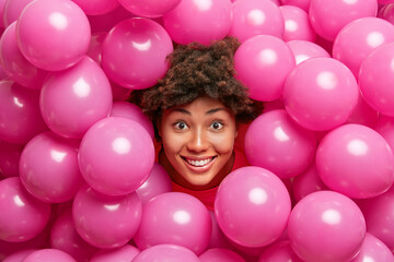 Fototapeta na wymiar Pretty cheerful young woman with Afro hair celebrates festive event has excited winsome expression surrounded by many inflated pink balloons has happy birthday meets guests. Holiday decoration