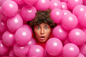 Fototapeta na wymiar Nice looking surprised dark skinned woman stares with great surprisement at camera sticks out head through inflated pink balloons shocked how many guests came on party. Amazed ethnic birthday girl