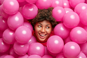 Fototapeta na wymiar Horizontal shot of happy curly haired adult woman smiles joyfully and looks away celebrates graduation party poses among inflated air balloons cheers over holiday has fun. Celebration concept
