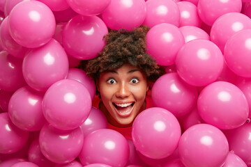 Fototapeta na wymiar Photo of postive crazy dark skinned woman looks happily at camera gets surprise has fun during festive day surrounded by small inflated rosy balloons. Female model prepares for housewarming party