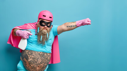 Horizontal shot of self confident superhero man pretends flying does chores about house poses with...