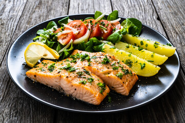 Fried salmon fillet with boiled potatoes, lemon and vegetable salad served on black plate on wooden table

