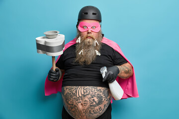 Household hygiene and cleaning service concept. Embarrassed bearded man housekeeper holds cleaning detergent and plunger dressed in superhero costume does house work isolated on blue background.