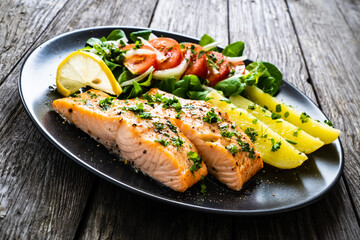 Fried salmon fillet with boiled potatoes, lemon and vegetable salad served on black plate on wooden table
