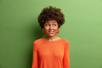 Horizontal shot of pretty curly haired young woman dressed in orange jumper bites lips and looks...